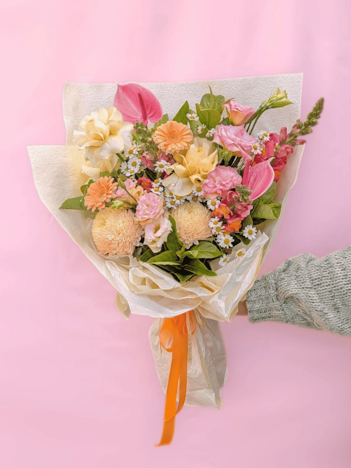 A hand wrapped in a light gray knit sweater holds a bouquet of pastel flowers wrapped in cream tissue paper with an orange ribbon, set against a solid pink background.
