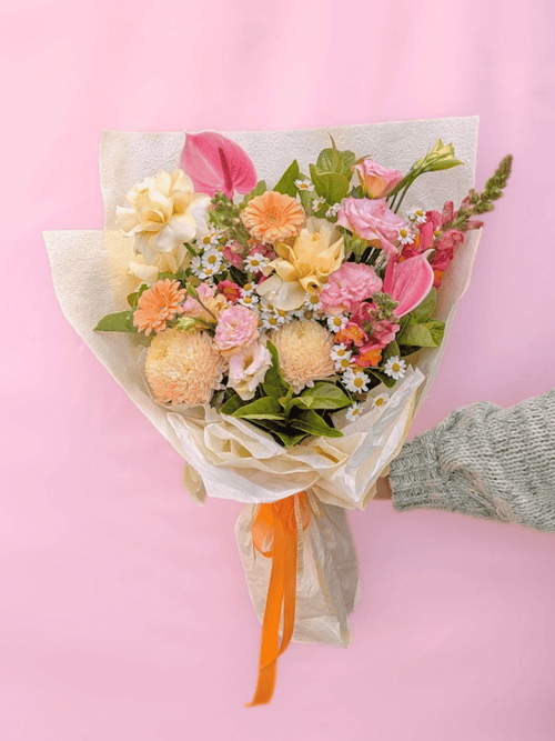 A hand wrapped in a light gray knit sweater holds a bouquet of pastel flowers wrapped in cream tissue paper with an orange ribbon, set against a solid pink background.