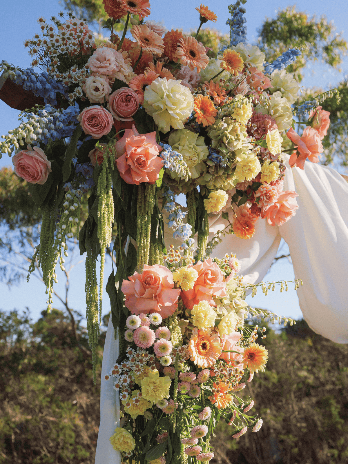 A large floral arch arrangement with peach, pink, cream, yellow, and blue flowers, green foliage, and white draped fabric, photographed outdoors against trees and blue sky.