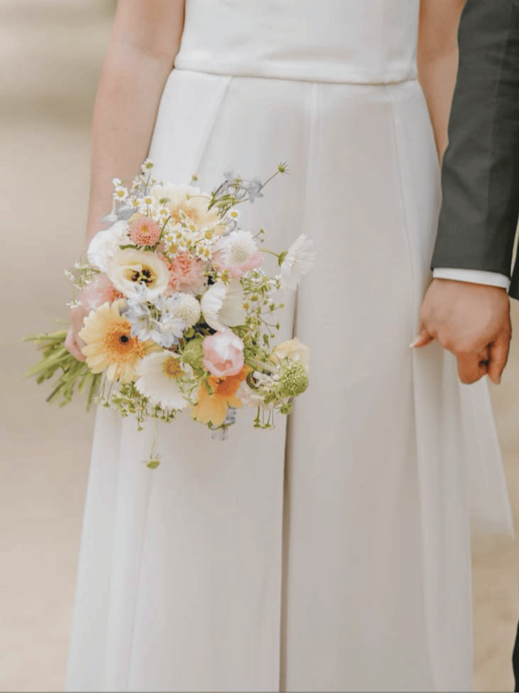 A close-up of a bride in a white wedding dress holding a pastel floral bouquet, with part of a groom's dark suit visible at the right edge.