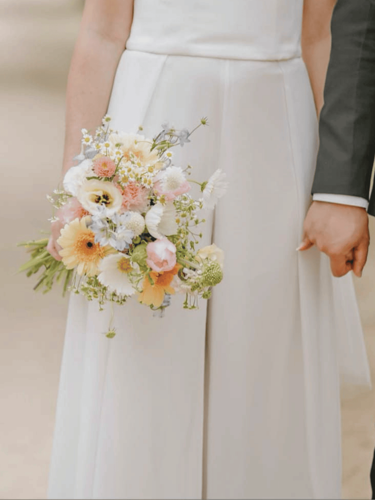 A close-up of a bride in a white wedding dress holding a pastel floral bouquet, with part of a groom's dark suit visible at the right edge.