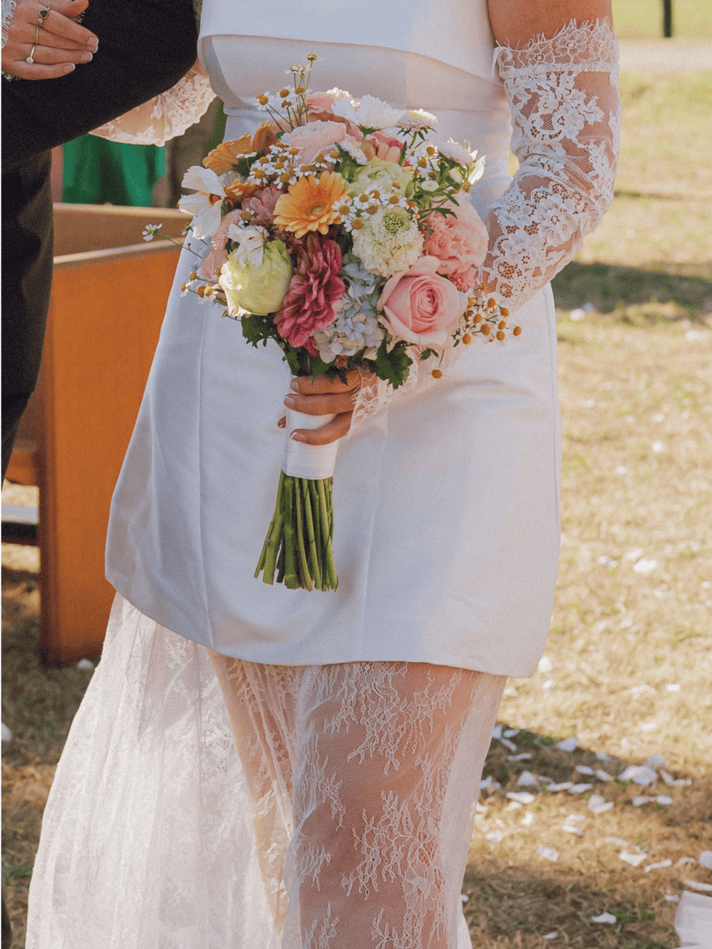 A close-up of a bride in a white satin wedding dress with long lace sleeves holding a colorful floral bouquet outdoors.