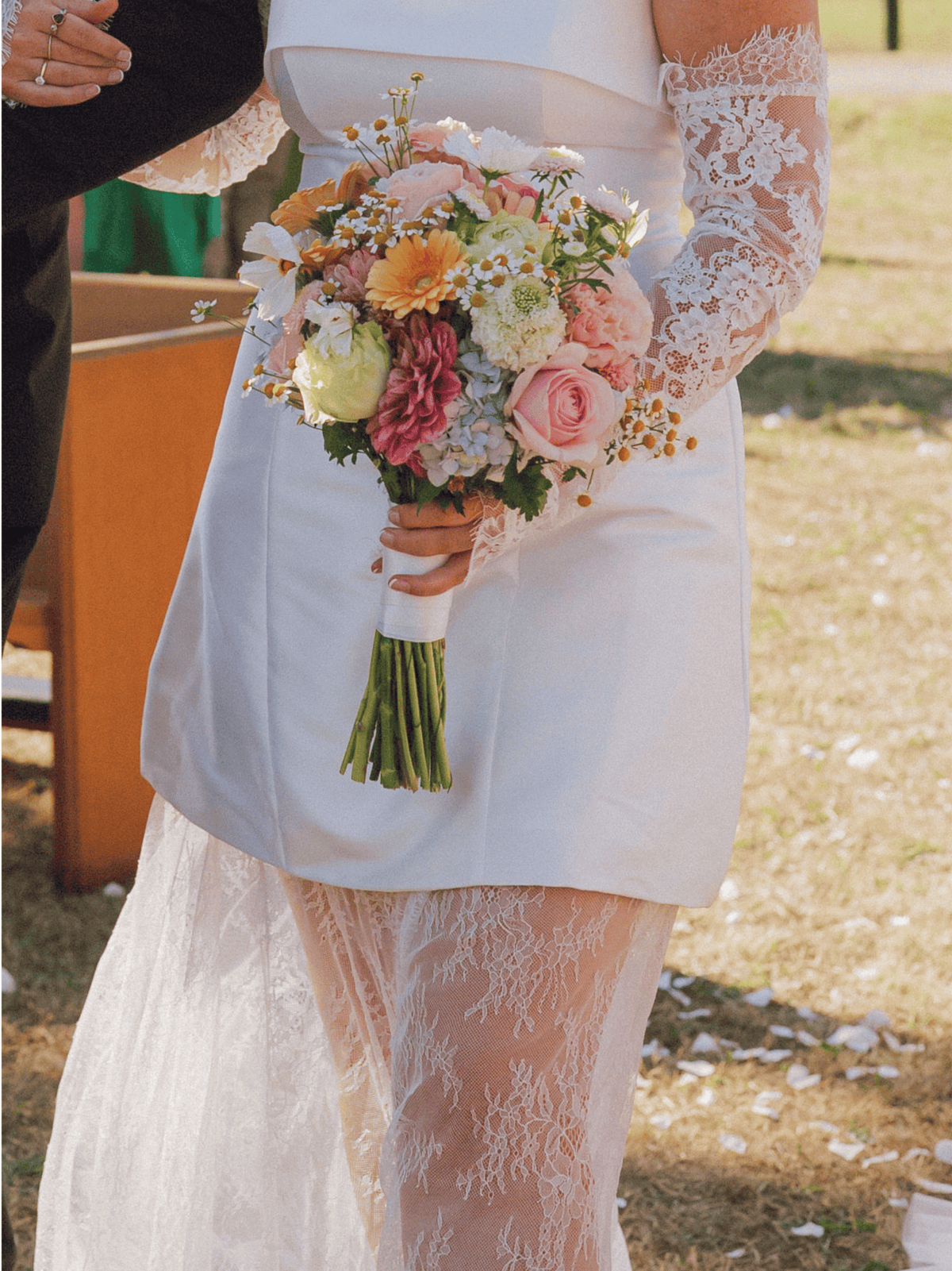 A close-up of a bride in a white satin wedding dress with long lace sleeves holding a colorful floral bouquet outdoors.