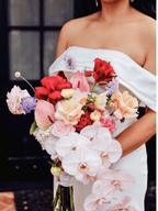 A close-up of a woman in a white off-the-shoulder dress holding a large, vibrant bridal bouquet filled with pink, red, peach, and white flowers.