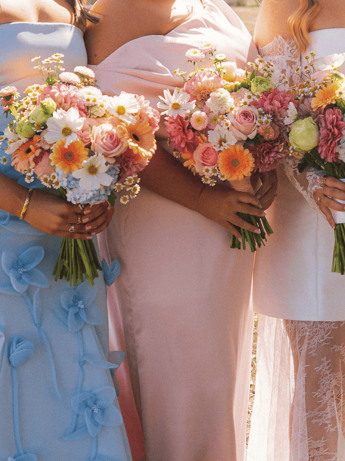 Three bridesmaids in pastel dresses holding vibrant floral bouquets outdoors. The dress on the left is light blue with 3D flower appliqués, the center dress is soft blush pink, and the dress on the right is white with lace detailing.