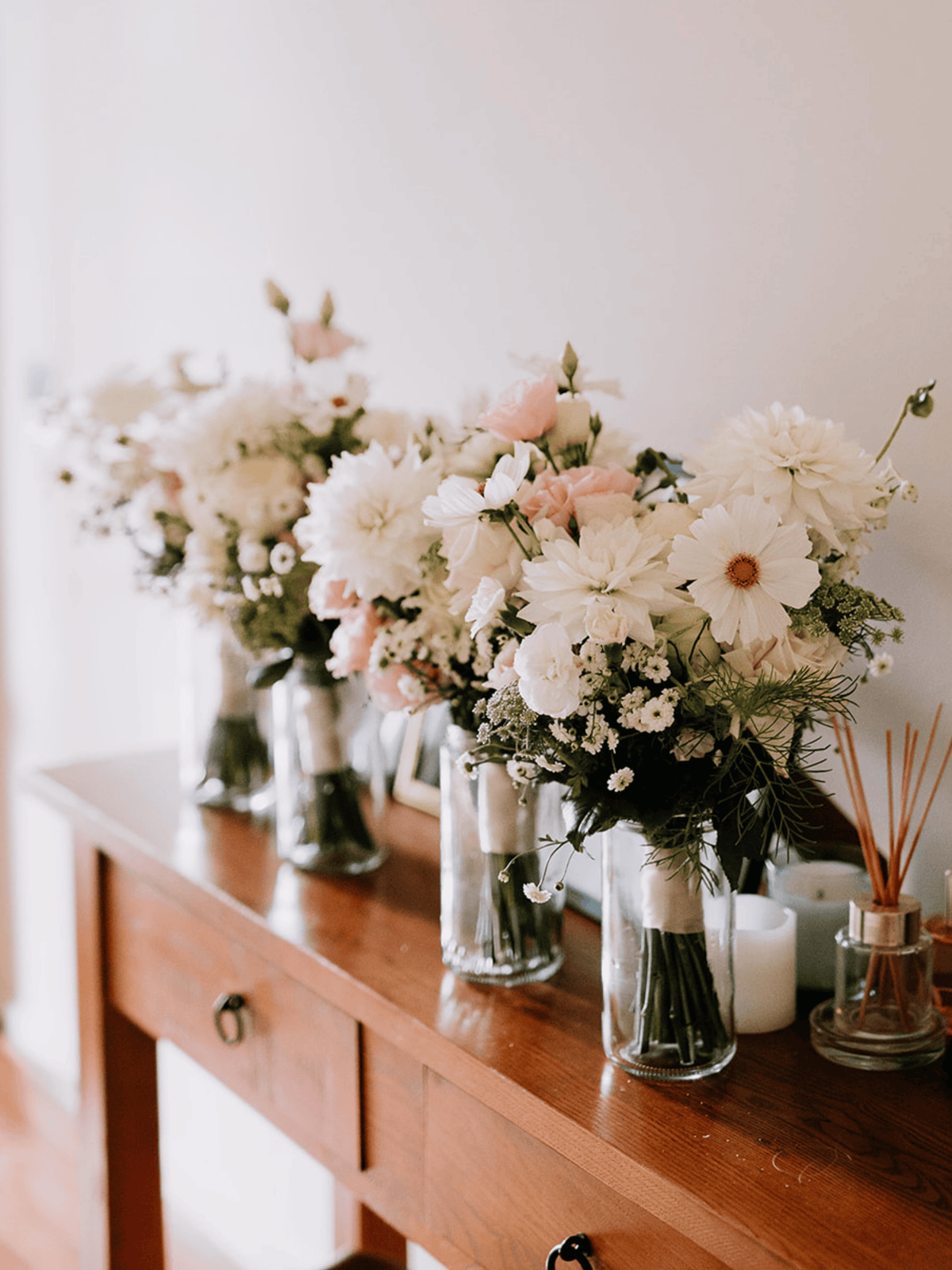 Three clear glass vases filled with white and blush pink floral arrangements sit on a wooden console table against a soft off-white wall.