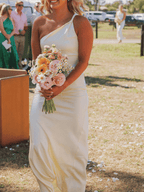 A woman outdoors wearing a one-shoulder cream satin gown and holding a pastel flower bouquet, with guests and a sunny outdoor setting blurred in the background.