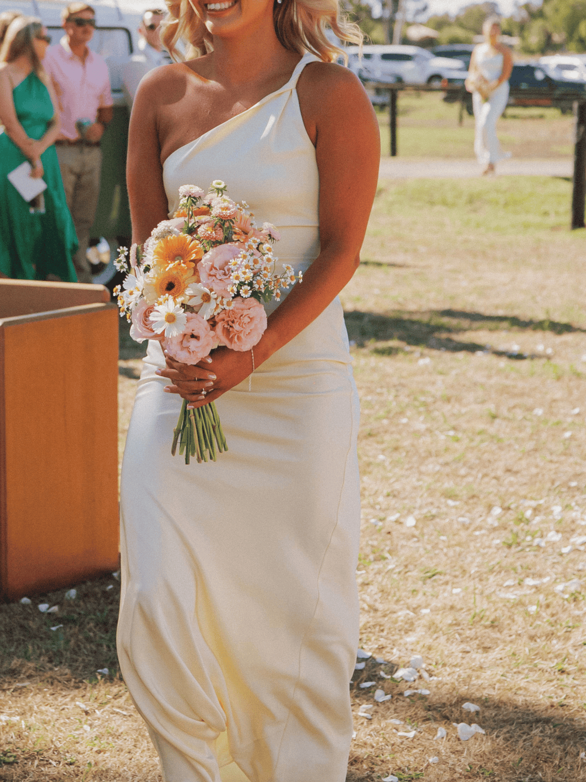 A woman outdoors wearing a one-shoulder cream satin gown and holding a pastel flower bouquet, with guests and a sunny outdoor setting blurred in the background.