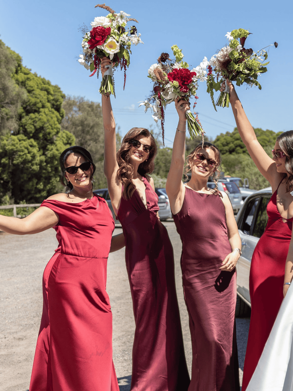 Four women outdoors wearing matching sleeveless bridesmaid dresses in shades of red, burgundy, and plum, each holding up a colorful bouquet under a blue sky with trees and parked cars in the background.