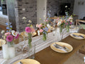 A long wooden dining table set for multiple guests with yellow plates, clear drinking glasses, white candles, and multiple floral centerpieces in glass vases, photographed in a real home interior with a brick column and kitchen visible in the background.