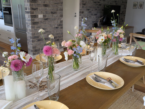 A long wooden dining table set for multiple guests with yellow plates, clear drinking glasses, white candles, and multiple floral centerpieces in glass vases, photographed in a real home interior with a brick column and kitchen visible in the background.
