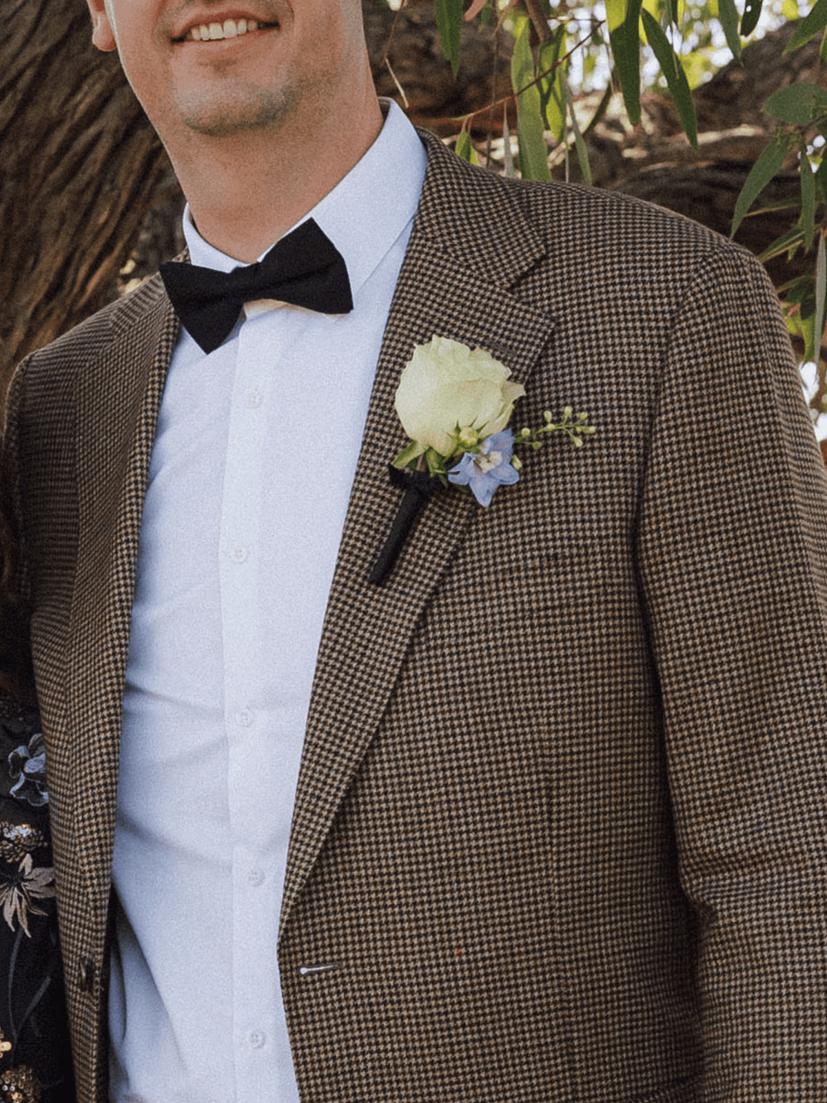 A man wearing a brown and black micro-check patterned suit jacket, a white dress shirt, a black bow tie, and a white rose boutonniere with a small blue flower, photographed outdoors in front of a tree.