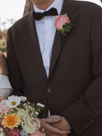 A close-up of a man wearing a dark brown textured suit jacket, white dress shirt, and black bow tie, with a pink rose boutonniere. He is holding hands with a woman holding a colorful bouquet of flowers.