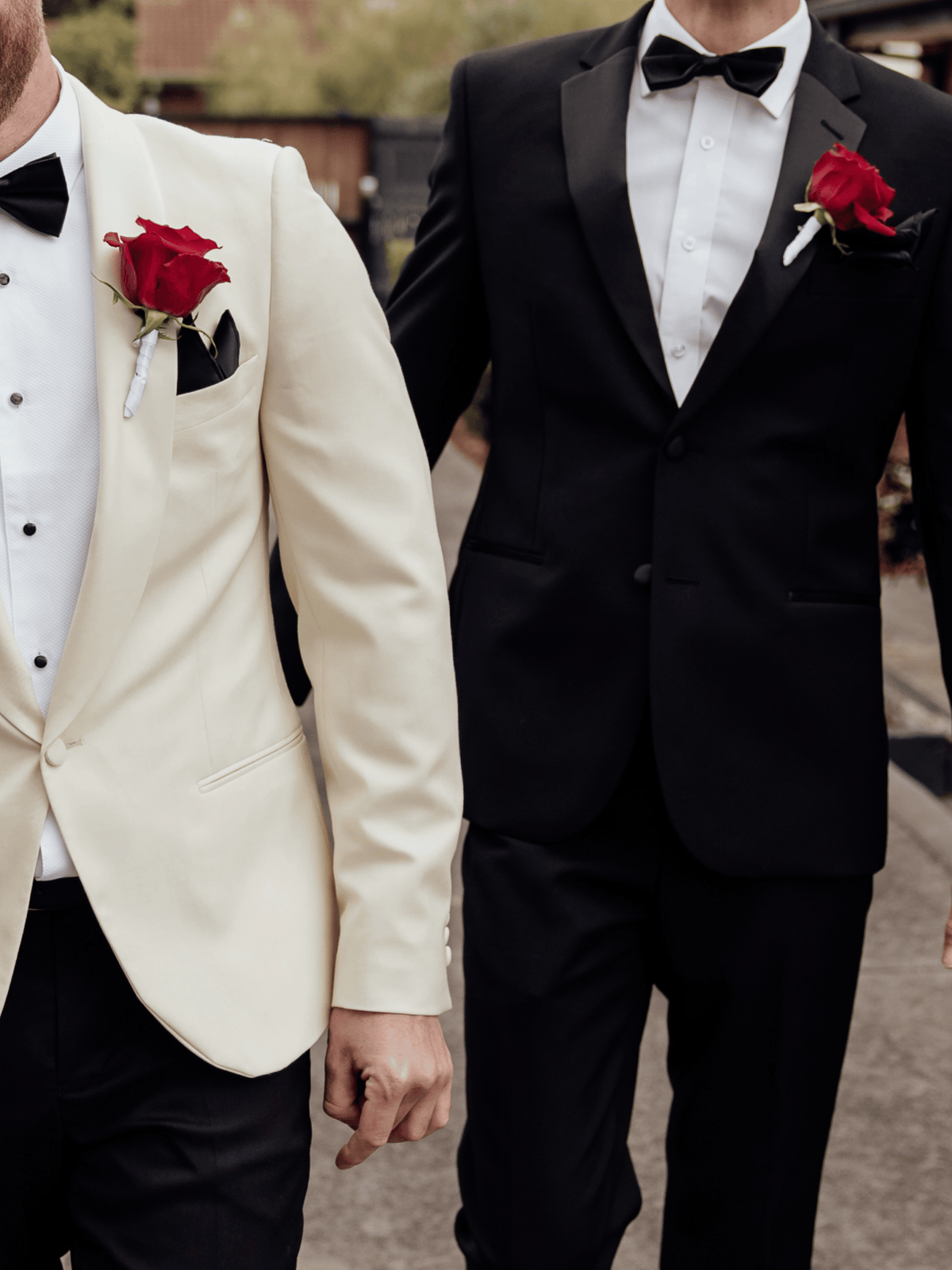 Two men in formal evening wear walking side by side, one wearing a cream tuxedo jacket with a red rose boutonniere and black trousers, and the other wearing a black tuxedo jacket with a red rose boutonniere and black trousers.