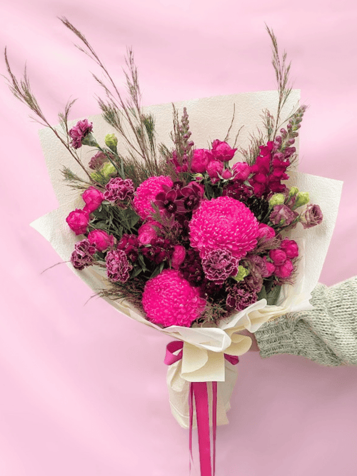 A vibrant bouquet of pink flowers, including bright pink chrysanthemums and darker magenta wildflowers, wrapped in cream paper with a pink ribbon, held by a hand wearing a green knit sleeve against a solid pink background.