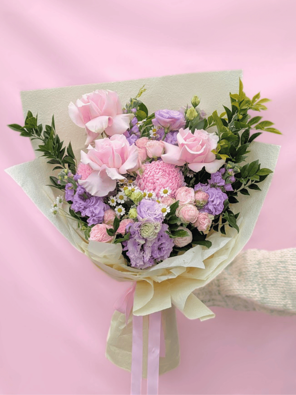 A pastel flower bouquet wrapped in cream paper with a pale pink ribbon, featuring pink roses, lavender blooms, green foliage, and small white daisies, held against a soft pink background.