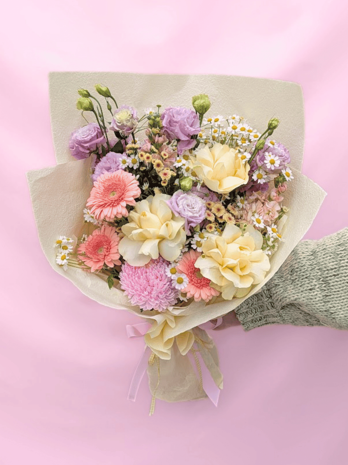 A pastel flower bouquet wrapped in beige paper, held by a hand wearing a gray knit sleeve, set against a solid pink background. The bouquet features cream carnations, pink and purple roses, purple lisianthus, and white daisies.