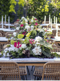 A long outdoor dining table decorated with a lush floral centerpiece featuring white, pink, and green flowers, set with woven rattan chairs and white taper candles in a garden setting.
