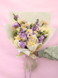 A hand-held bouquet of pastel flowers wrapped in cream paper with a ribbon, held by a hand wearing a green knit sleeve against a solid pink background.