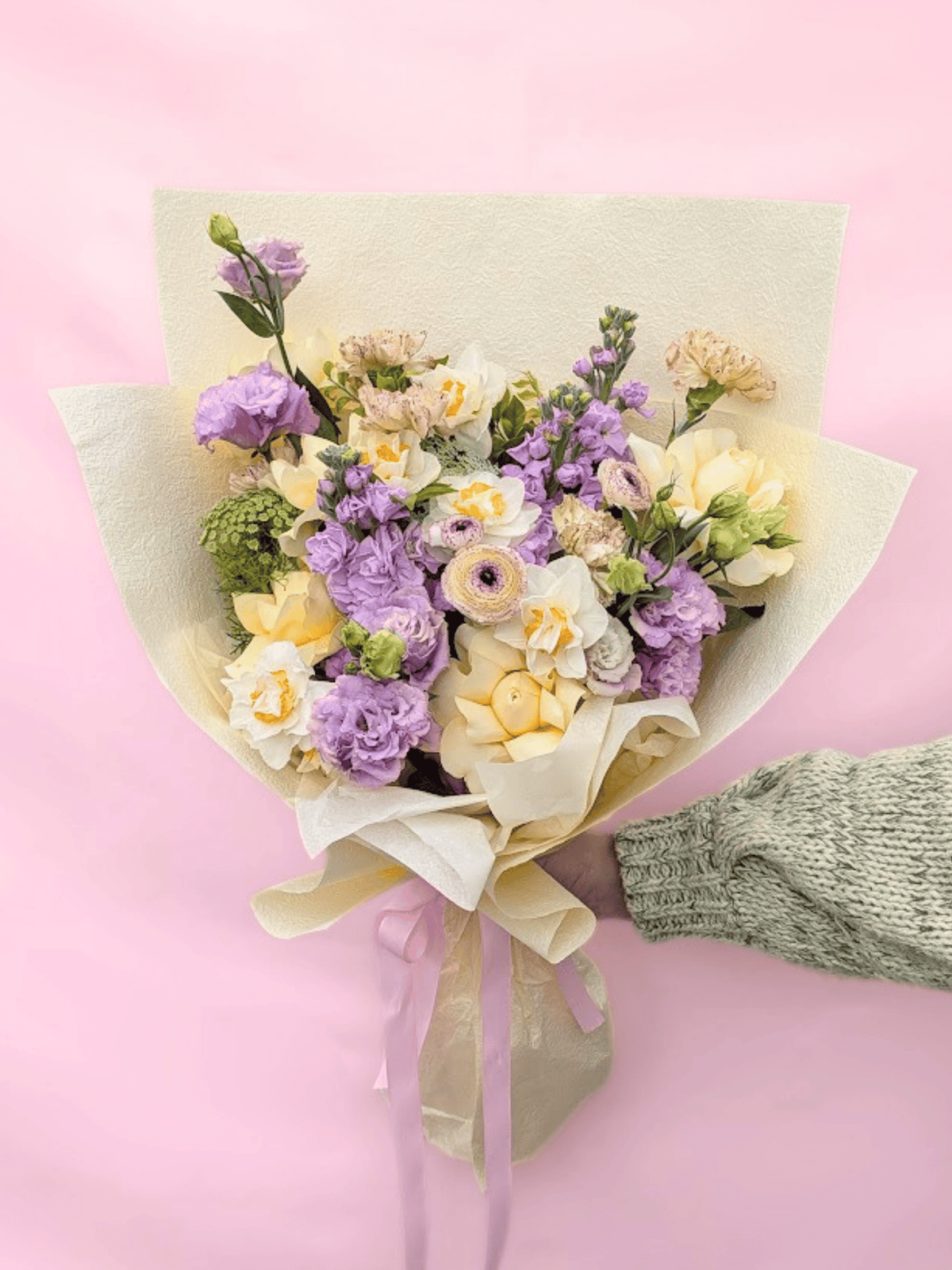 A hand-held bouquet of pastel flowers wrapped in cream paper with a ribbon, held by a hand wearing a green knit sleeve against a solid pink background.