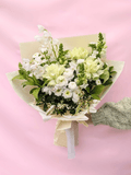 A bouquet of white and pale yellow flowers with green foliage, wrapped in cream paper and tied with a white ribbon, held by a hand wearing a textured green sleeve against a solid pink background.