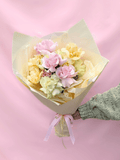 A bouquet of pastel pink, pale yellow, and light green roses wrapped in cream paper with a pink ribbon, held by a hand wearing a green knit sleeve against a solid pink background.