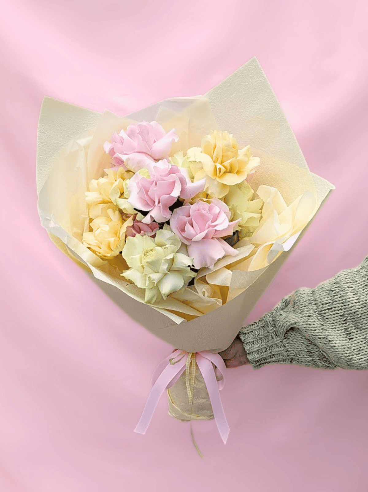 A bouquet of pastel pink, pale yellow, and light green roses wrapped in cream paper with a pink ribbon, held by a hand wearing a green knit sleeve against a solid pink background.