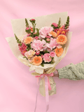 A hand wrapped in a light green knit sleeve holds a small bouquet of pink and orange flowers wrapped in cream paper with a pink ribbon, set against a solid pink background.
