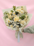 A bouquet of cream and white flowers with green foliage, wrapped in beige tissue paper and held by a hand wearing a gray knit sweater against a solid pink background.