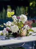 A rustic floral centerpiece with white roses, green foliage, and purple wildflowers arranged on a white wooden table outdoors.