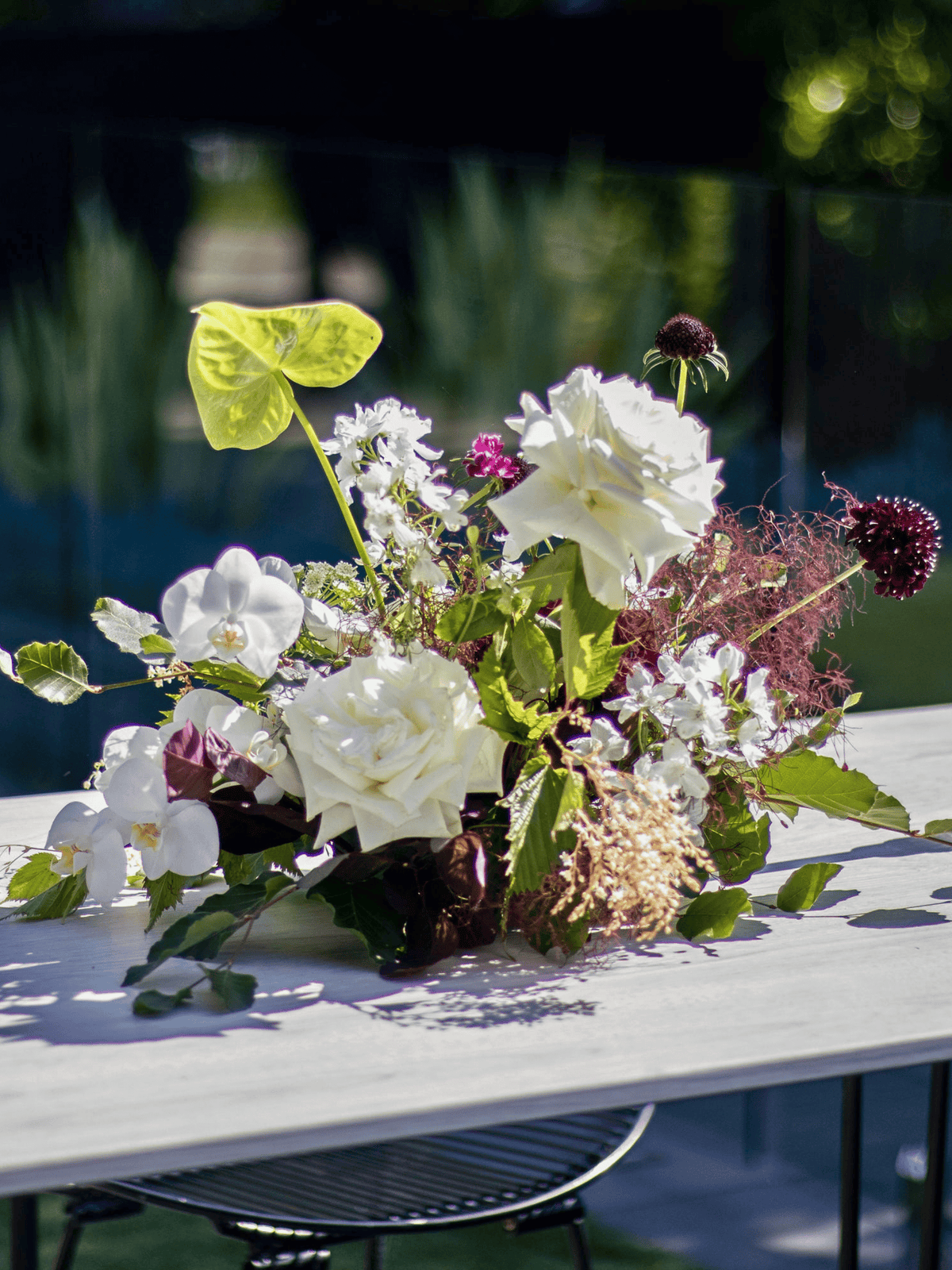 A rustic floral centerpiece with white roses, green foliage, and purple wildflowers arranged on a white wooden table outdoors.