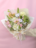 A pastel flower bouquet wrapped in cream paper and tied with a pale pink ribbon, held by a hand wearing a beige knit sleeve against a soft pink background.
