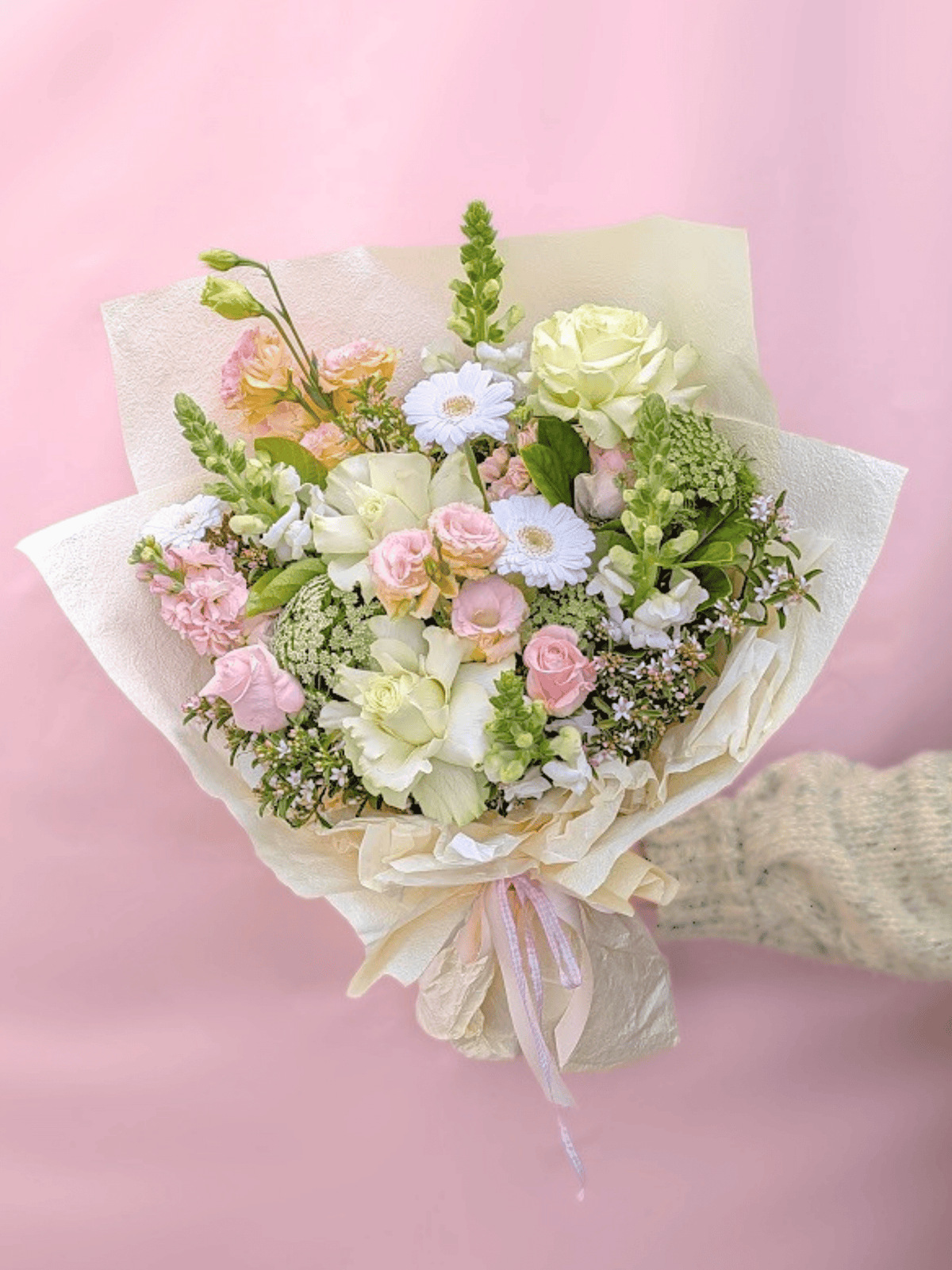 A pastel flower bouquet wrapped in cream paper and tied with a pale pink ribbon, held by a hand wearing a beige knit sleeve against a soft pink background.