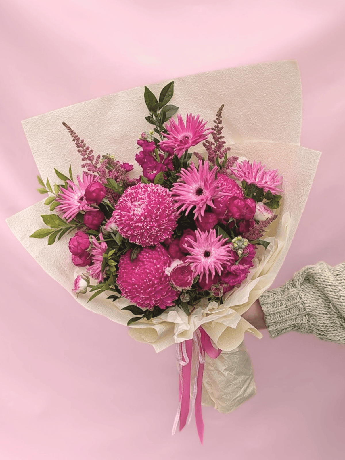 A vibrant bouquet of pink flowers, including gerbera daisies and mums, wrapped in textured cream paper with pink ribbons, held by a hand wearing a green knit sweater against a solid pink background.