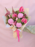 A vibrant pink flower bouquet wrapped in beige paper and tied with a pink ribbon, held by a hand wearing a green knit sleeve against a soft pink background.
