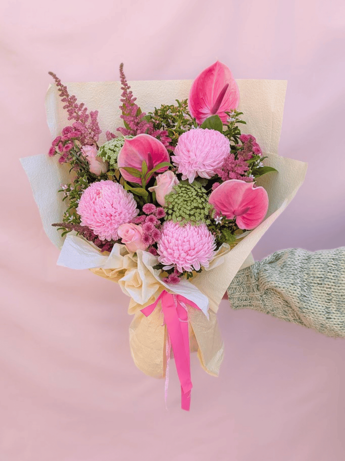 A vibrant pink flower bouquet wrapped in beige paper and tied with a pink ribbon, held by a hand wearing a green knit sleeve against a soft pink background.
