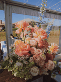 A large, vibrant floral arrangement featuring peach roses, pink carnations, white daisies, and blue delphiniums, displayed on a rustic wooden table under a clear-sided event tent.
