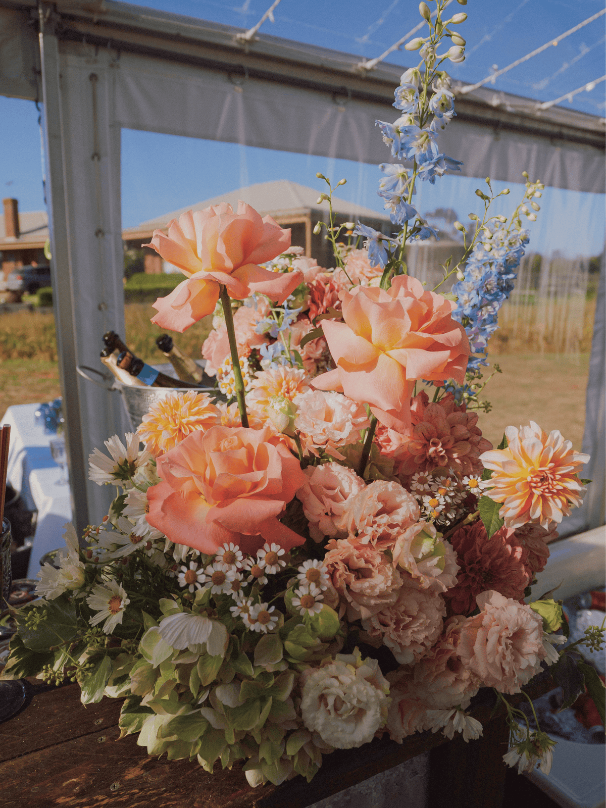 A large, vibrant floral arrangement featuring peach roses, pink carnations, white daisies, and blue delphiniums, displayed on a rustic wooden table under a clear-sided event tent.