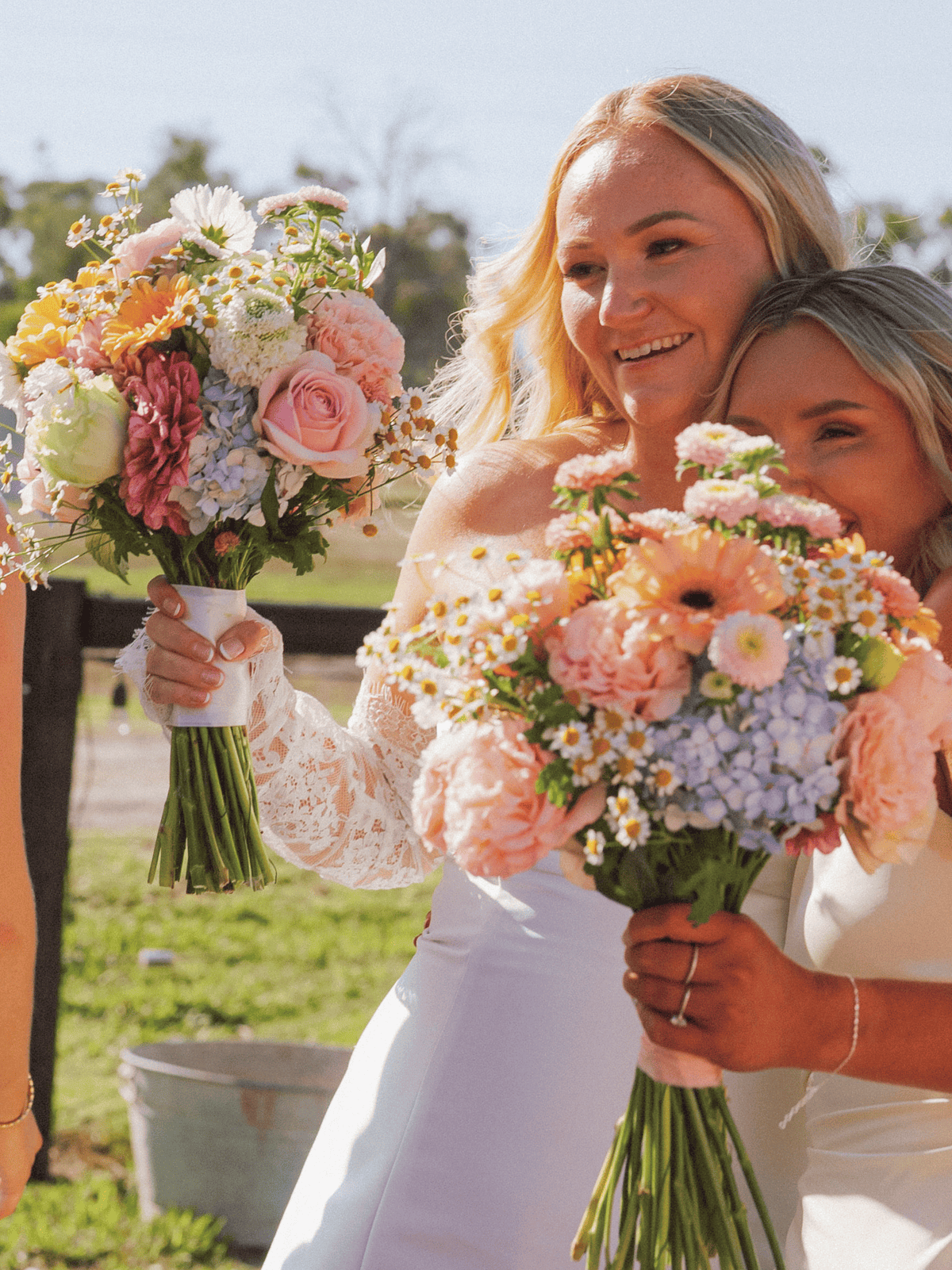 Two women outdoors holding large pastel flower bouquets. The woman in the foreground wears a white lace bridal dress and smiles while holding a bouquet with pink, peach, white, and light blue flowers.